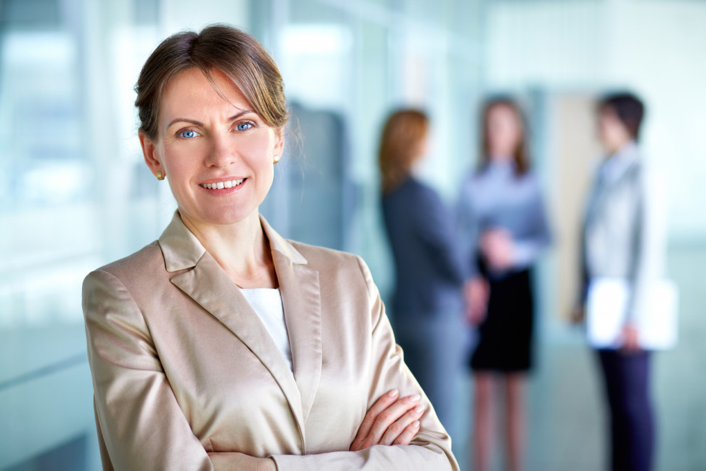 female boss smiling with arms crossed in office with employees in blurred background
